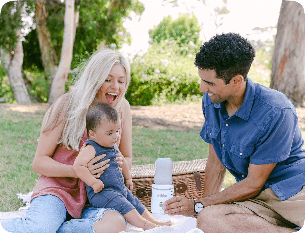 Smiling parents use the Babba portable baby bottle warmer at a picnic, keeping breast milk warm while traveling.